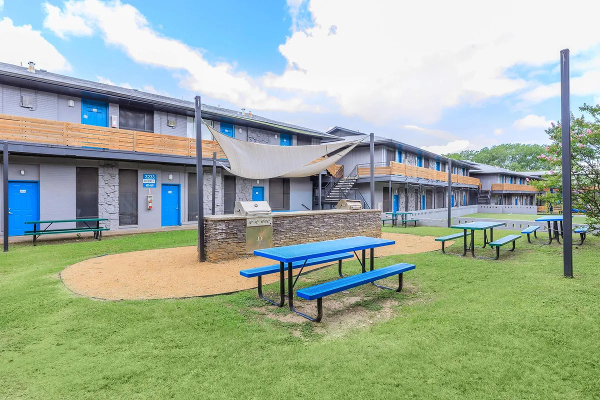 Shade sail over a stone grill station with blue picnic tables on a courtyard lawn between two-story buildings at 3311 at Vernon in Dallas