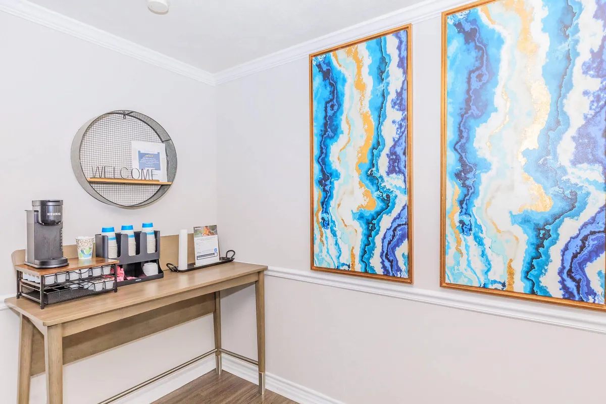 Coffee station with a single-serve brewer, cup dispenser, and framed abstract wall art in the leasing office at 3311 at Vernon in Dallas