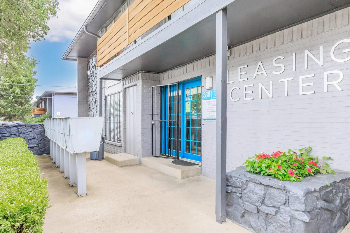 Leasing center entrance with a blue-framed glass door, wall-mounted mailboxes, and a stone planter with flowers at 3311 at Vernon in Dallas