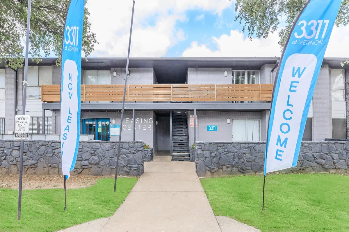 Leasing center exterior with a concrete walkway, stone half walls, wood balcony railing, and two vertical banner flags at 3311 at Vernon in Dallas