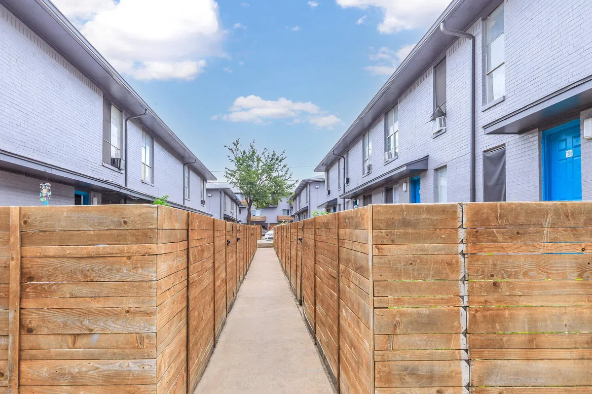 Concrete walkway between wood patio fences and two-story gray brick buildings with blue doors at 3311 at Vernon in Dallas