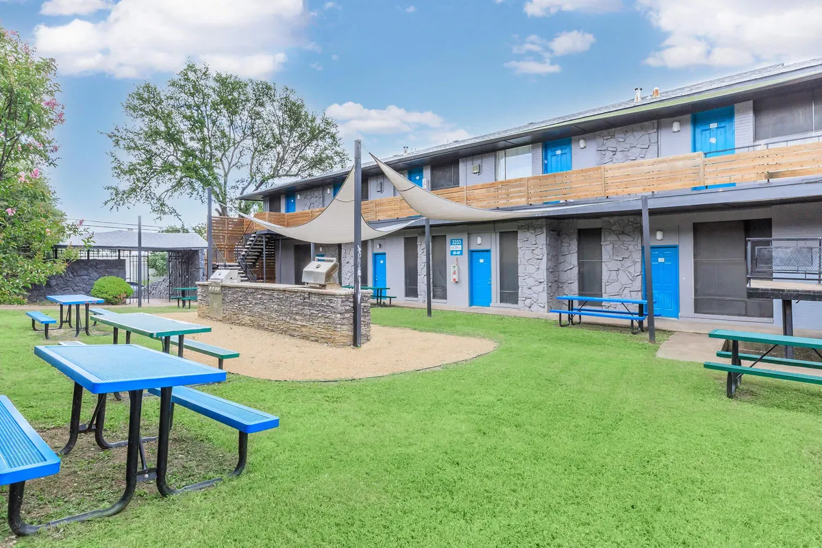Courtyard lawn with blue and green picnic tables, grills, and shade sails beside a two-story building with blue doors at 3311 at Vernon in Dallas