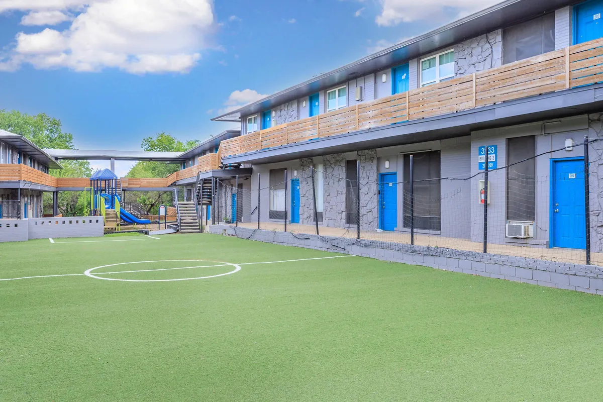 Turf soccer field along a two-story building with blue doors and wood balcony railings, with a playground slide in the background at 3311 at Vernon in Dallas