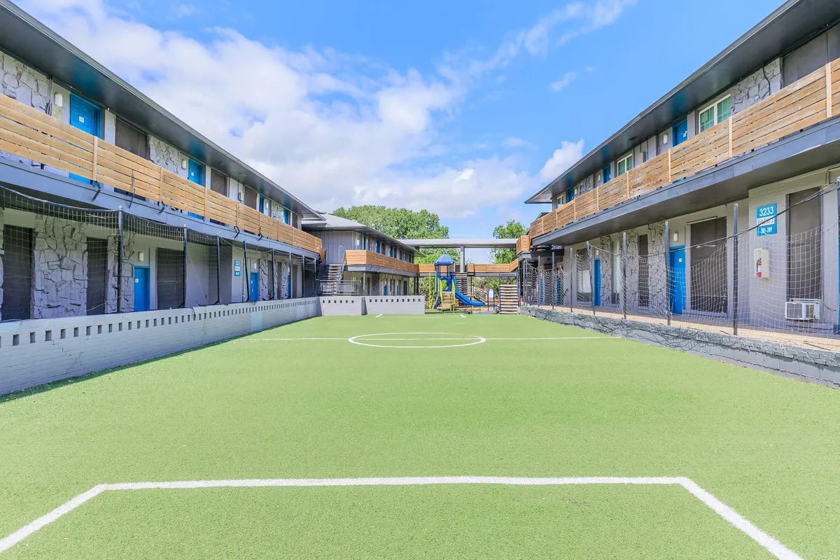 Turf soccer field with a center circle between two two-story buildings, with a playground at the far end at 3311 at Vernon in Dallas