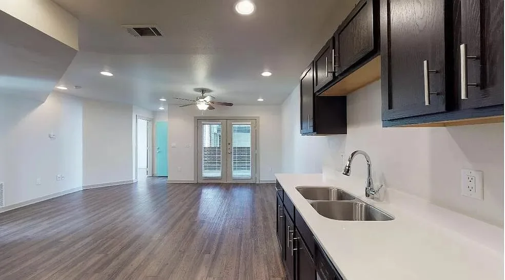 Open living space with a ceiling fan opening to a dark-cabinet kitchen and French doors to a patio