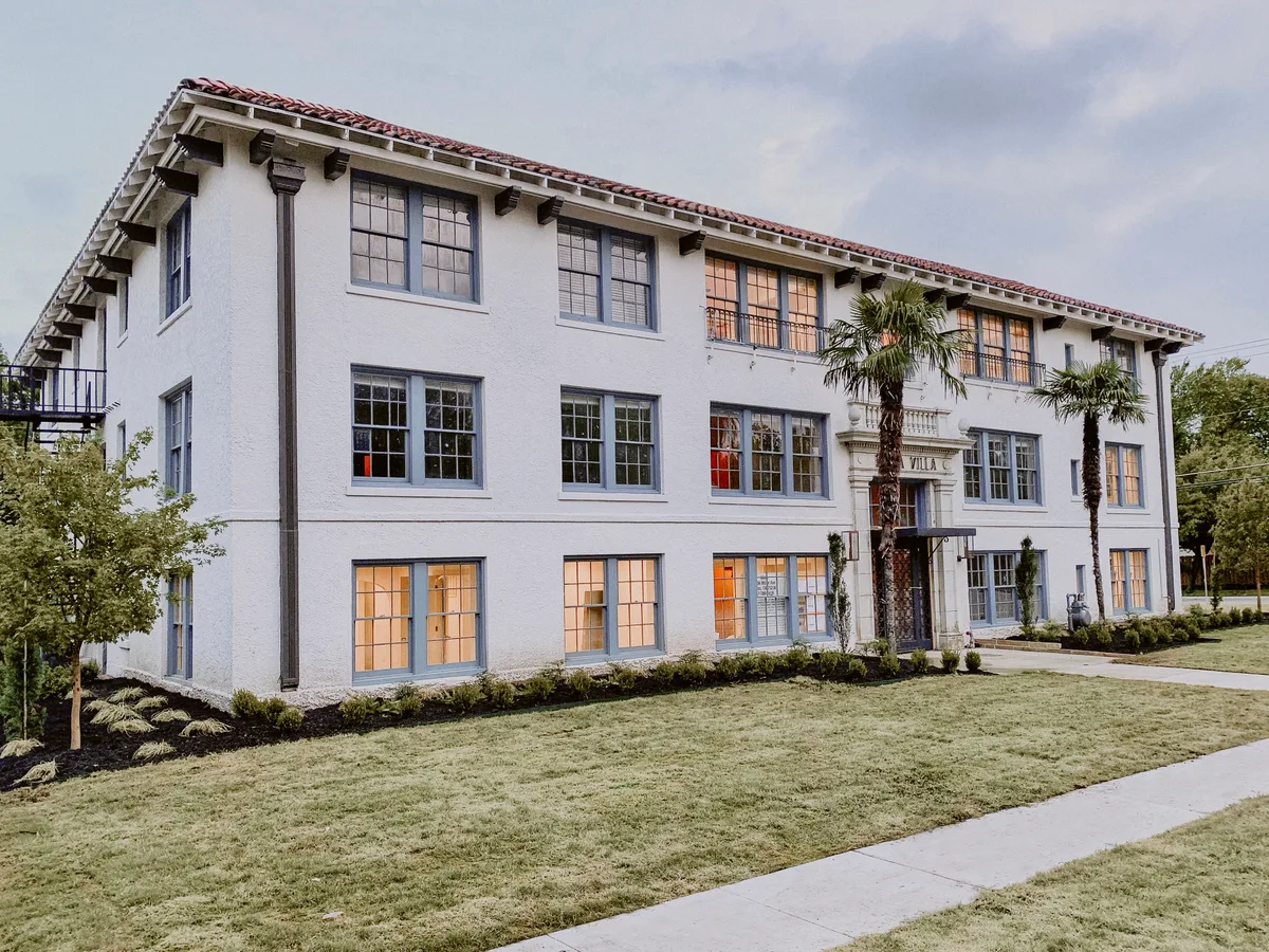 Three-story white stucco building at dusk with red barrel-tile roof, blue-gray window trim and warm-lit windows