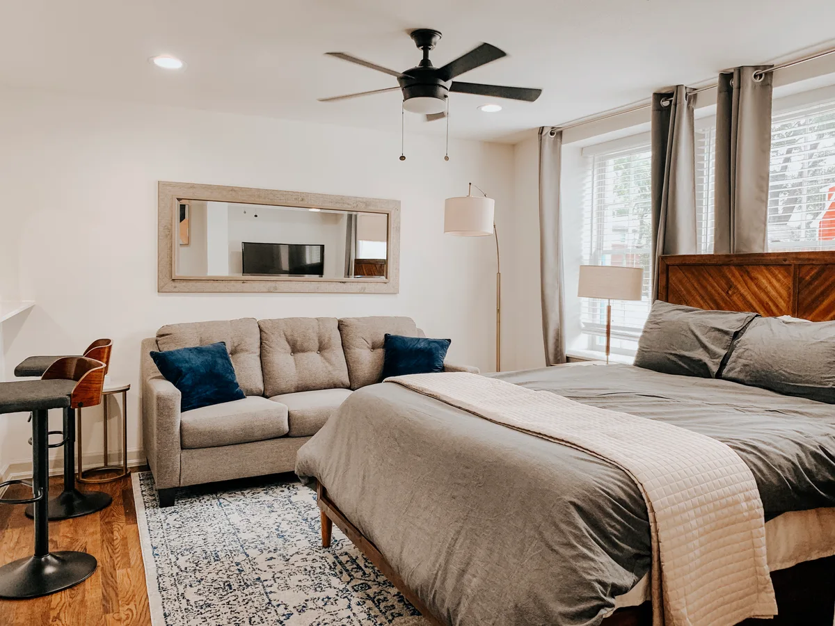 Studio sleeping and lounge area with bed, gray sofa and a framed mirror under a black ceiling fan