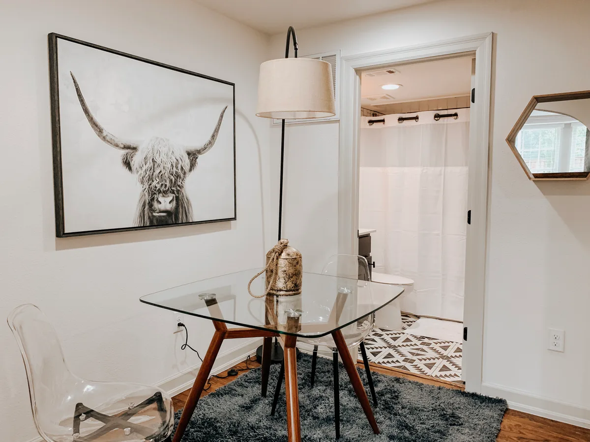 Dining corner with glass table, acrylic chairs and framed Highland-cow art beside the bathroom door
