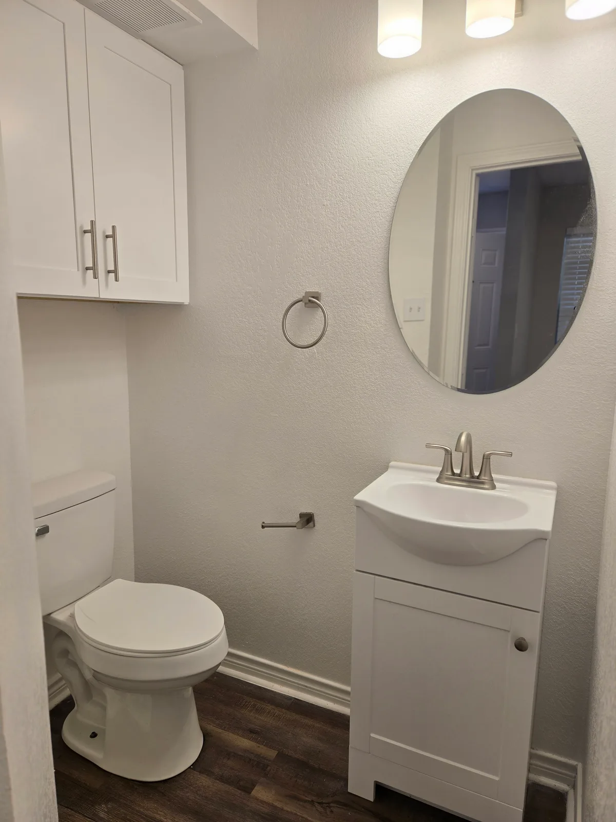 Bathroom with an oval mirror, a white single vanity, an over-toilet storage cabinet, and wood-look flooring