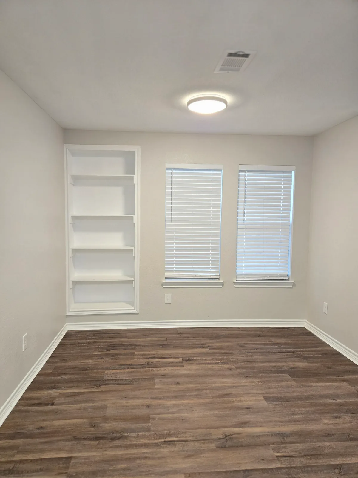 Bright room with recessed white built-in shelving, two blinded windows, and wood-look plank flooring