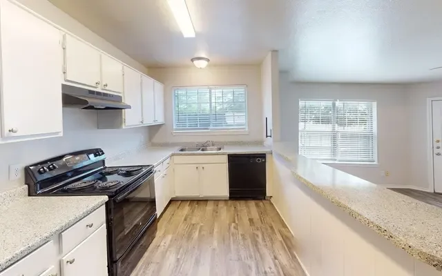 Galley kitchen with white cabinetry, black appliances, granite-look counters, and a window above the double sink