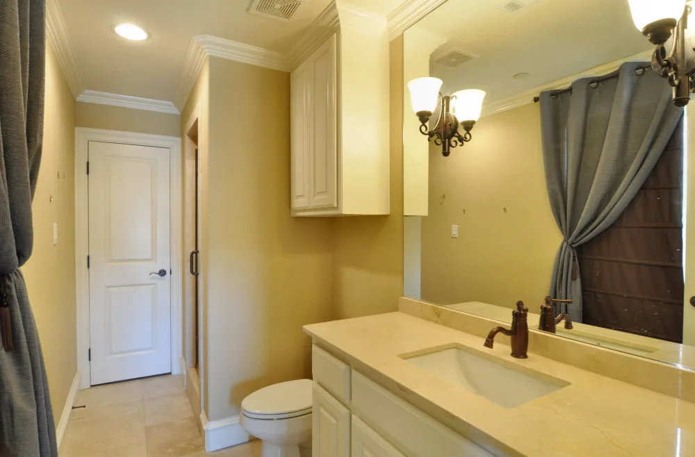 Bathroom with cream marble vanity, framed mirror, sconce lighting, toilet, and curtained window