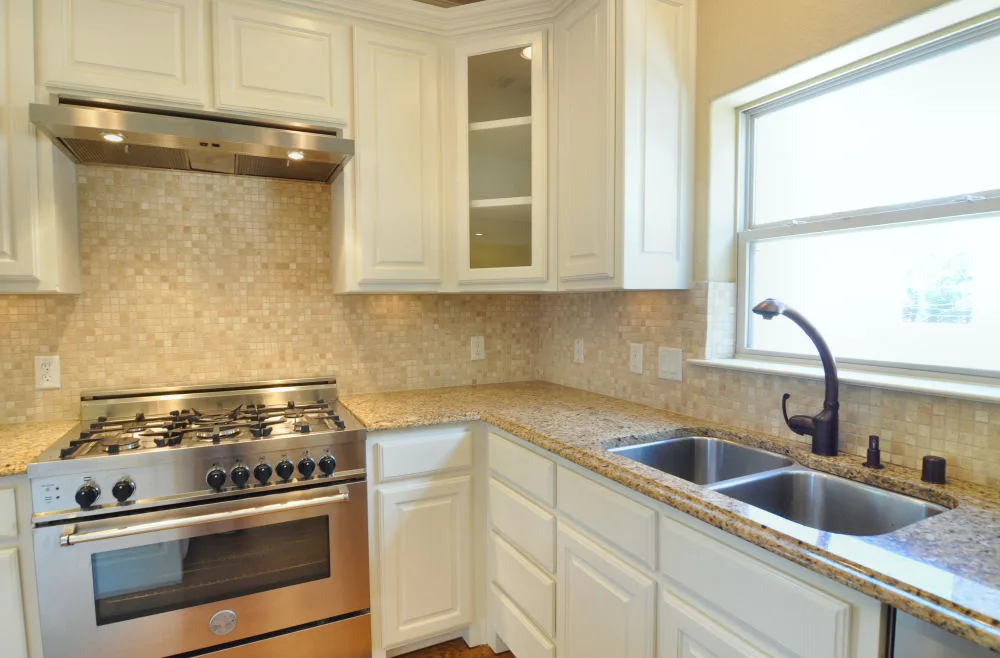 Stainless gas range with vent hood, mosaic backsplash, and granite counters beside a double sink