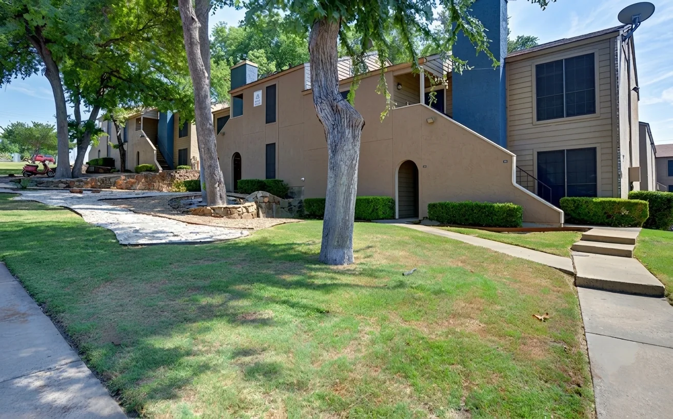Two-story garden-style apartment buildings with tan stucco and lap-siding exteriors, exterior stairwells, and mature shade trees over a landscaped, tree-lined lawn