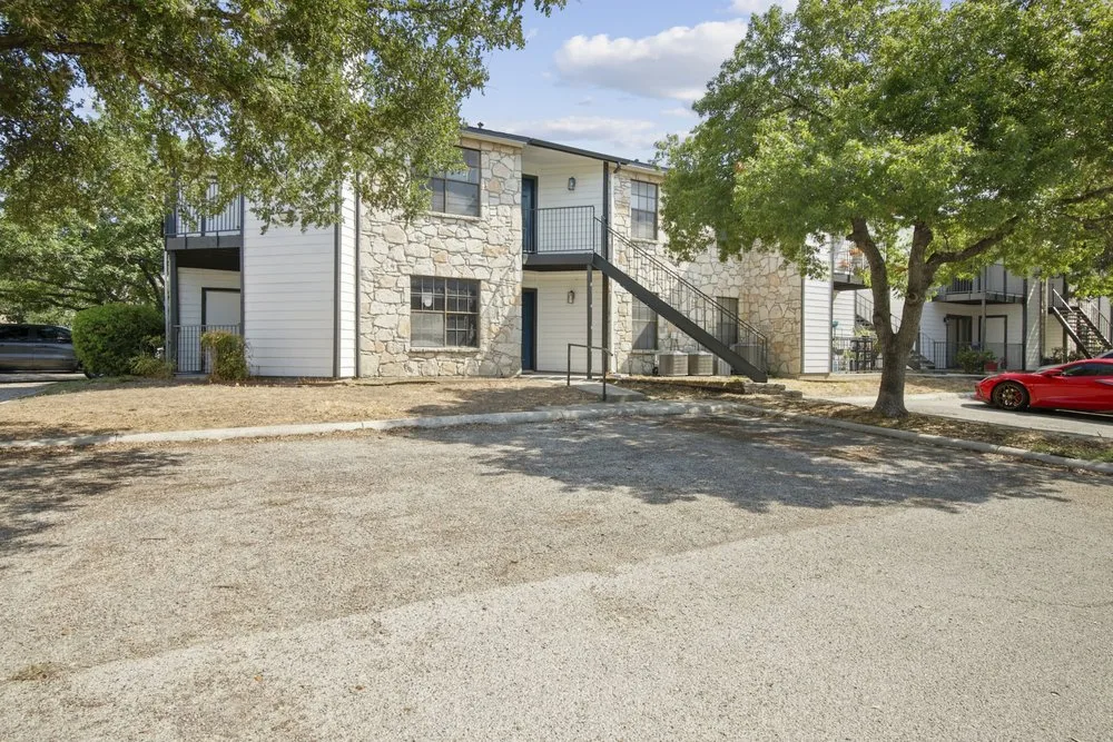 Two-story apartment building with white siding, natural stone, and an exterior staircase under mature shade trees