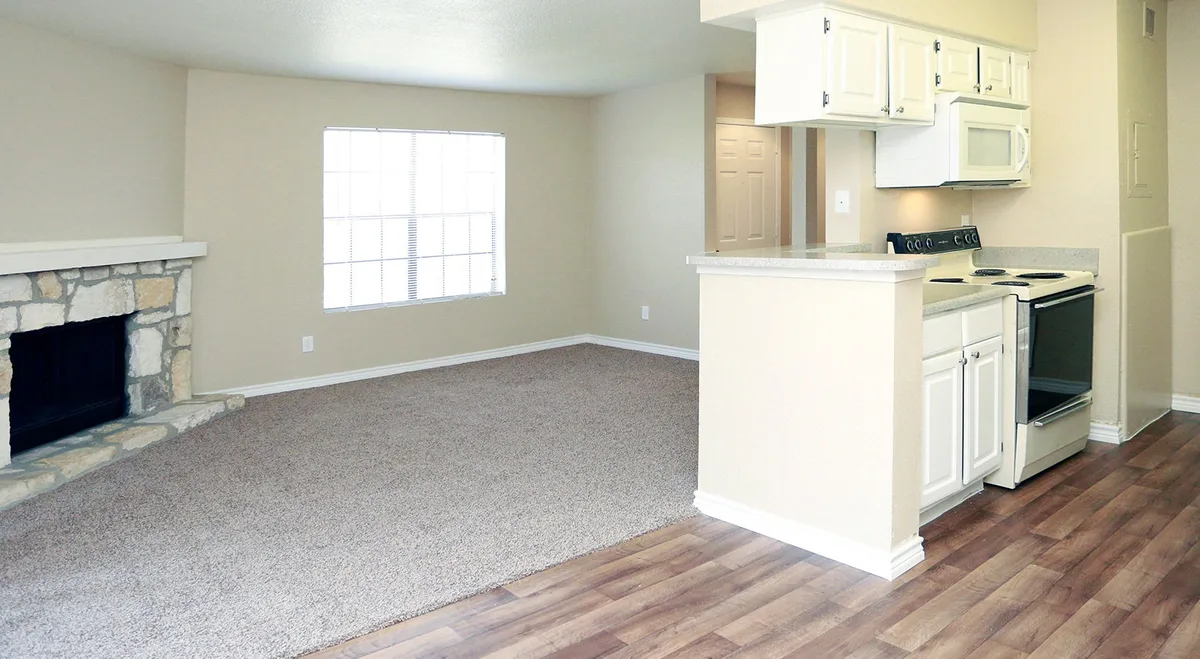 Open living room with a stone corner fireplace, plush carpet, wood-look entry flooring, and an adjoining white kitchen