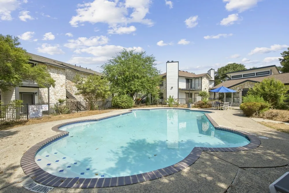 Kidney-shaped swimming pool with brick coping, a shaded patio table, and stone apartment buildings beyond