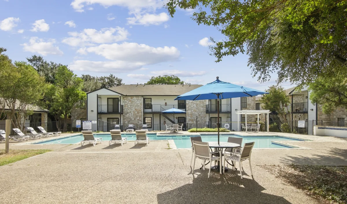 Sparkling community pool with cushioned lounge chairs, a shade umbrella, and a patio dining set framed by oak trees
