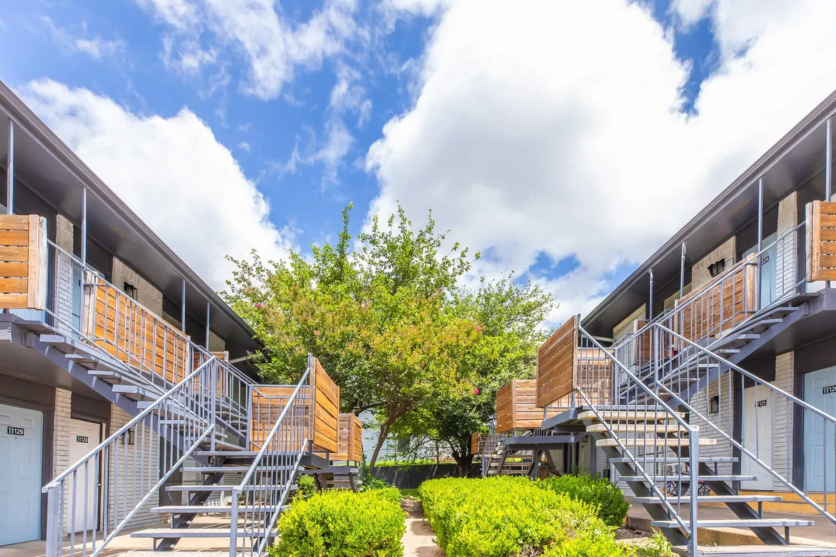 Courtyard with hedges between two-story buildings with wood-slat balcony screens and exterior stairs at La Madera in Dallas