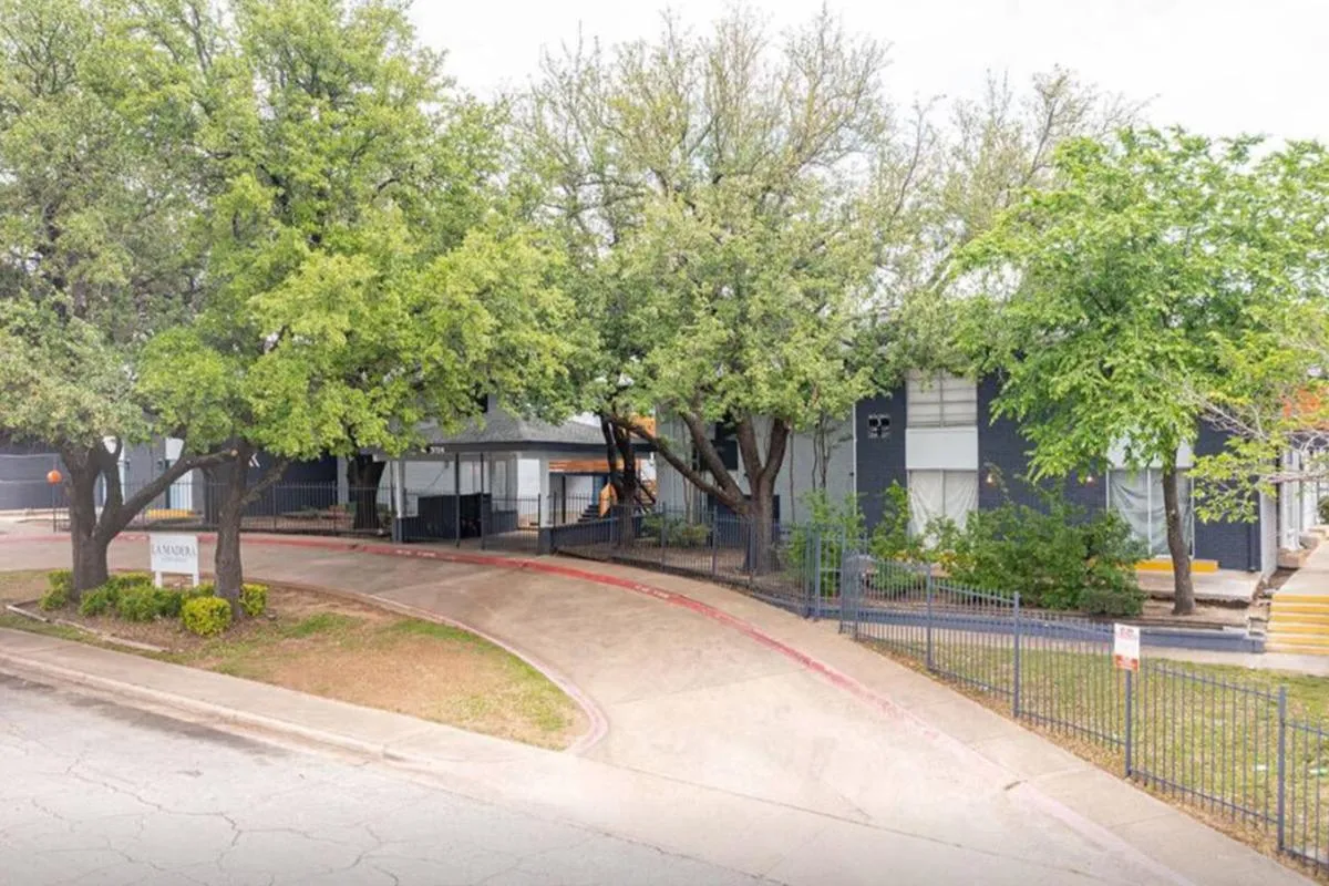 Street view of the curved entrance drive, iron fence, and shade trees at La Madera in Dallas