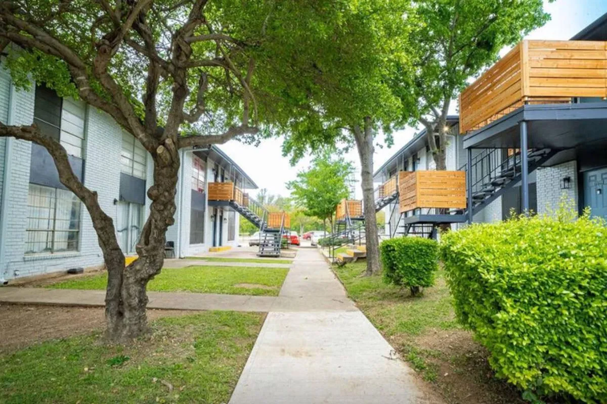 Sidewalk between two-story buildings with wood-slat balcony screens and shade trees at La Madera in Dallas