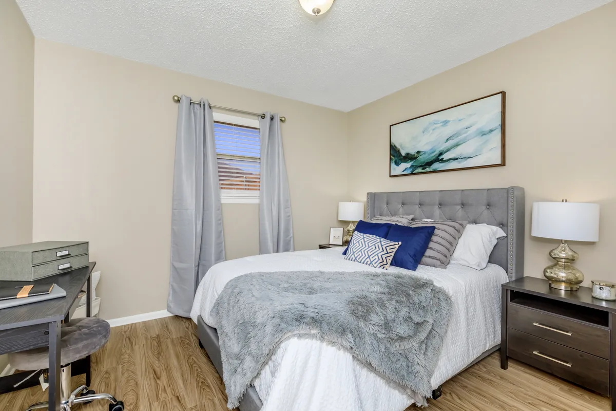 Staged bedroom with a gray upholstered bed, wood-look flooring, a desk nook, and a bright curtained window