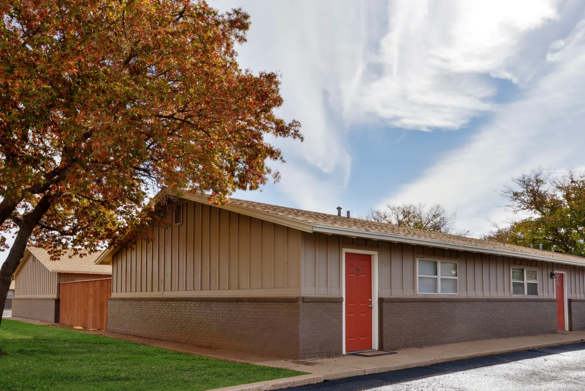 One-story building with tan board-and-batten siding, a brick base, and a red entry door beside an autumn oak tree