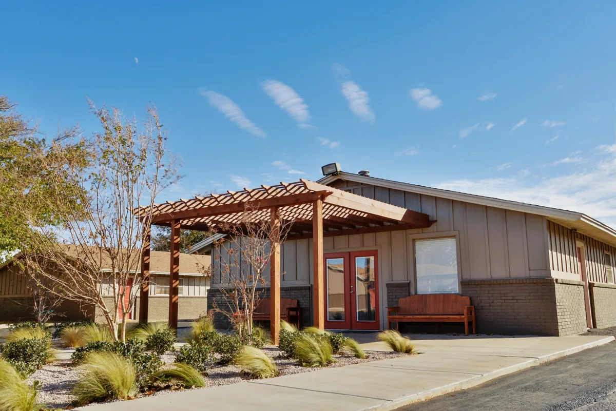 Oak Creek leasing office with a cedar pergola, red double doors, and xeriscaped grasses under a blue Lubbock sky