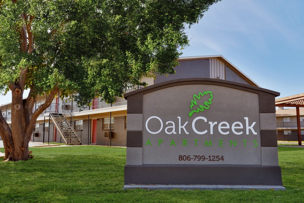 Oak Creek Apartments entrance monument sign with the leaf logo, set on a green lawn in front of the residential buildings