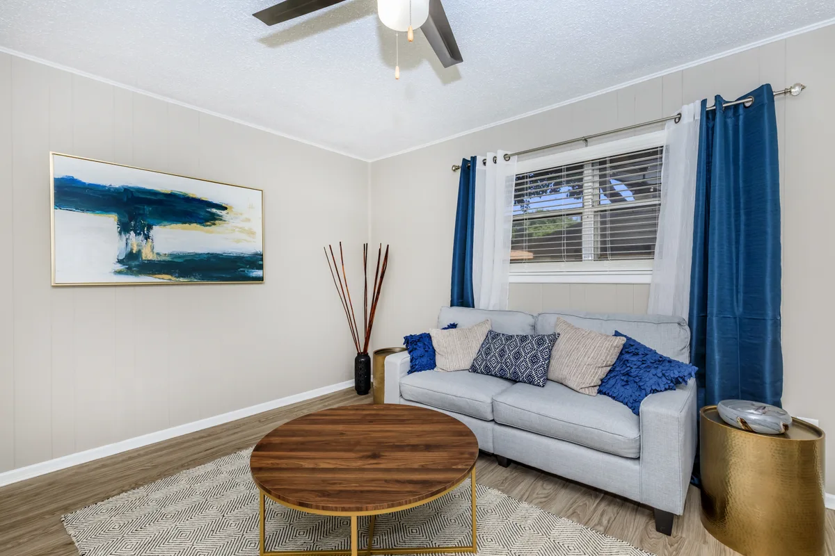 Bright living room with a gray sofa, blue accent pillows, framed abstract art, and a round wood coffee table