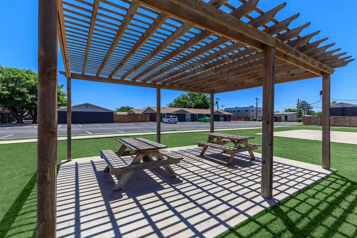 Wood pergola shading two picnic tables on a concrete pad framed by green lawn