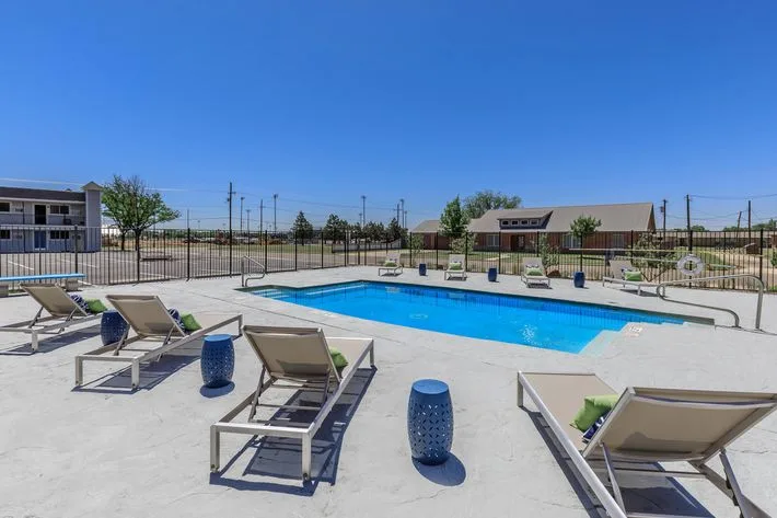 Fenced swimming pool with cushioned lounge chairs and blue accent stools beside the community clubhouse