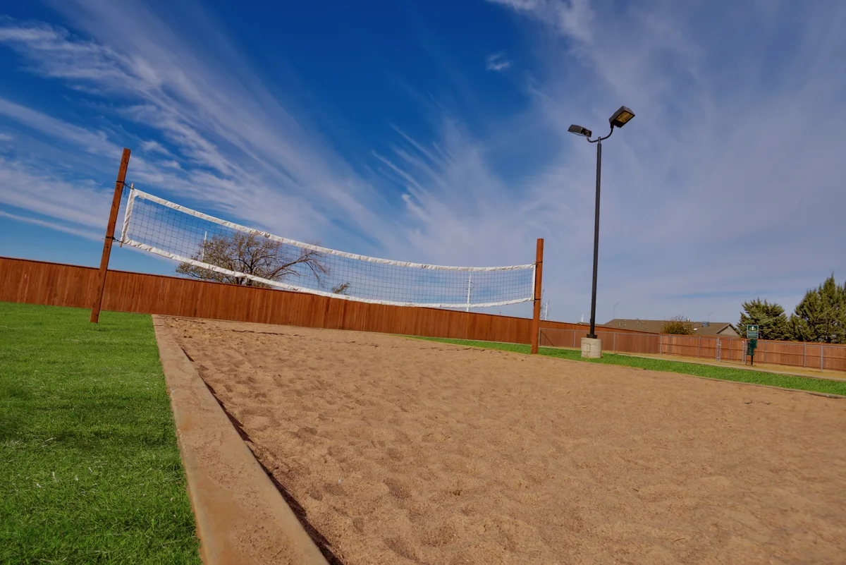 Sand volleyball court with a full net, wood privacy fencing, and a light pole under a bright blue sky