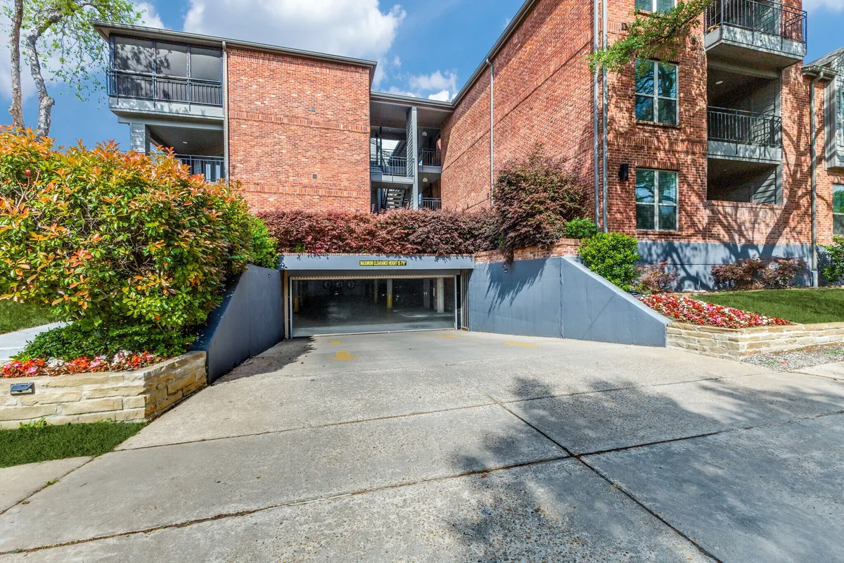 Covered parking garage entrance ramp flanked by flower beds and brick building