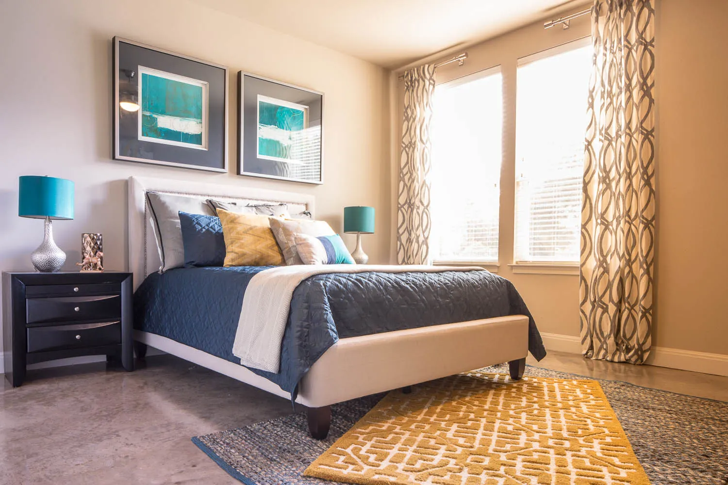 A staged bedroom with a queen bed, two windows and a patterned rug