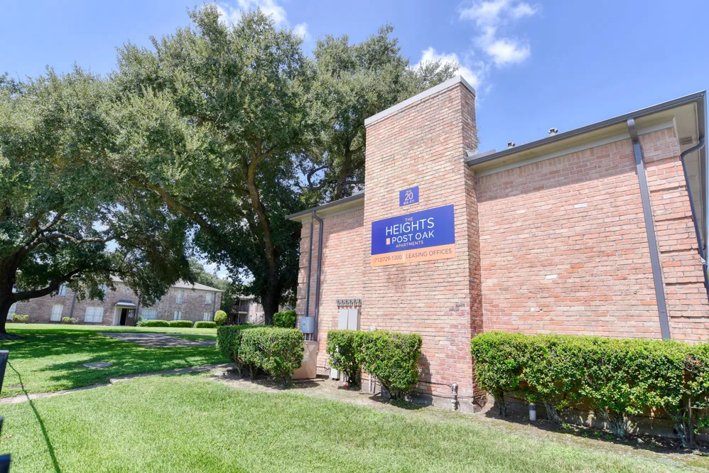 Red-brick apartment building with property signage, mature oak trees, and manicured lawn