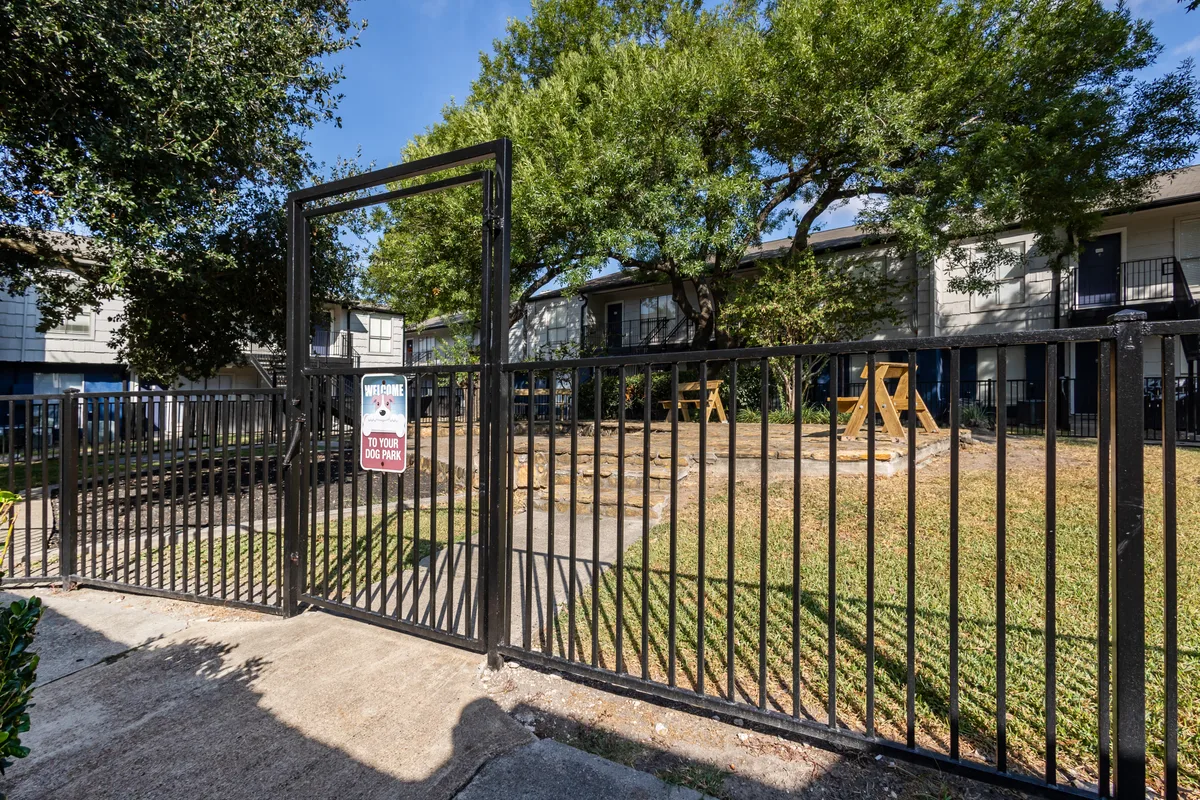 Gated on-site dog park with welcome sign, picnic tables, and shade trees