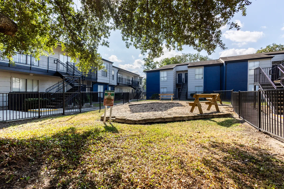 Fenced dog park with a picnic table and benches between residential buildings