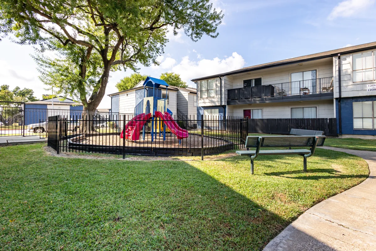 Children's playground with slides enclosed by fencing beside a shaded bench