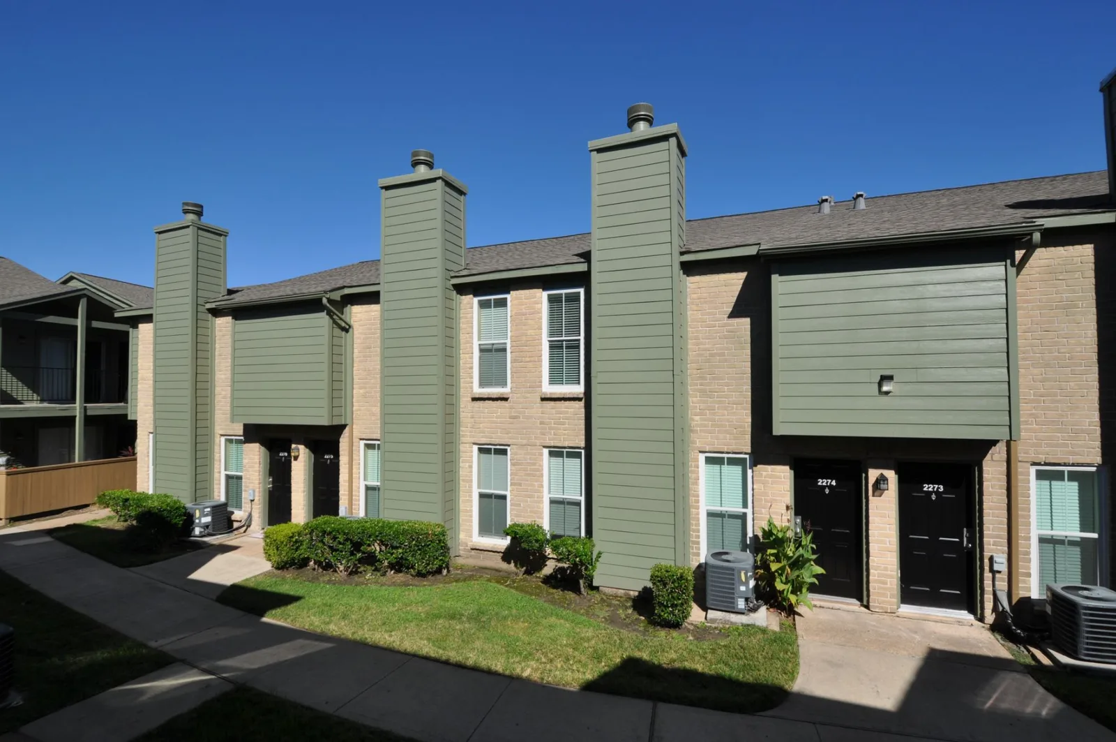 Two-story green-and-brick buildings with black entry doors along a landscaped walkway
