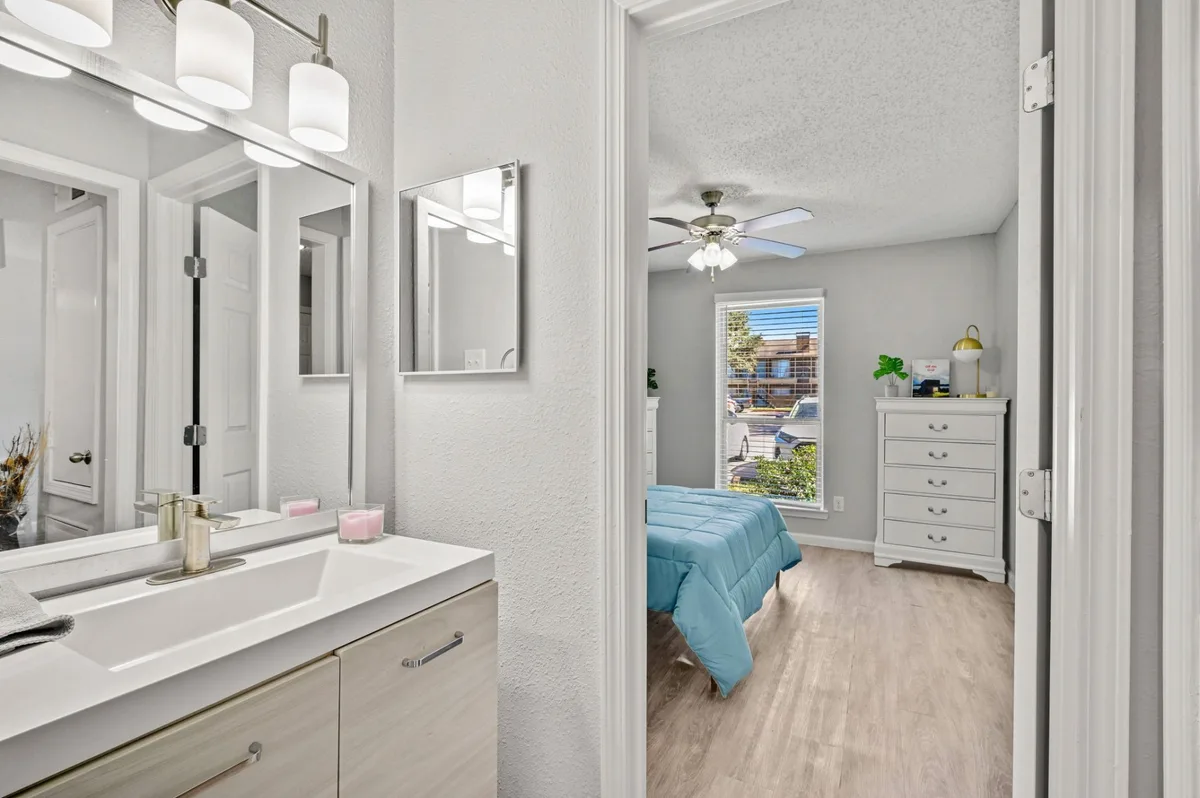 Bathroom vanity with a wood-tone cabinet and framed mirror beside an open bedroom
