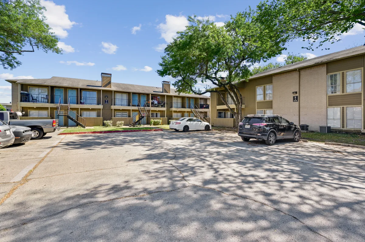 Two-story tan-brick garden apartment buildings behind a mature shade tree and open parking