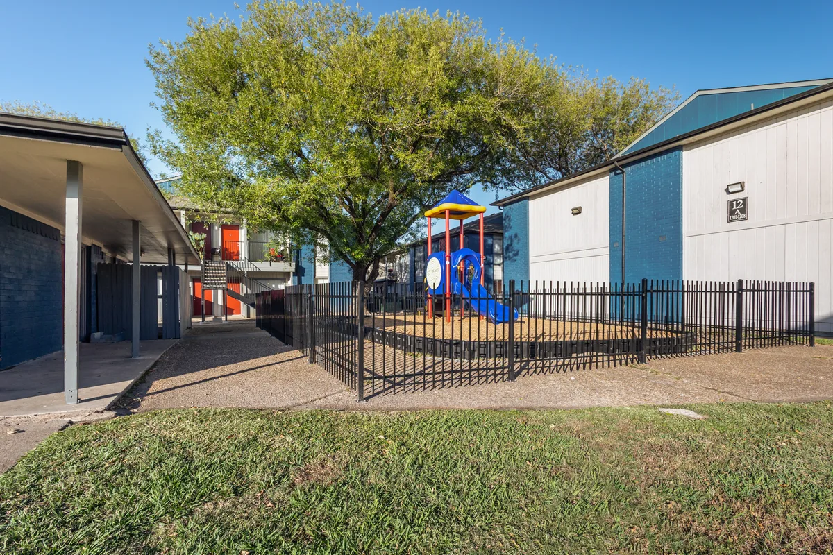 Outdoor playground enclosed by a black fence, surrounded by buildings and a grassy area