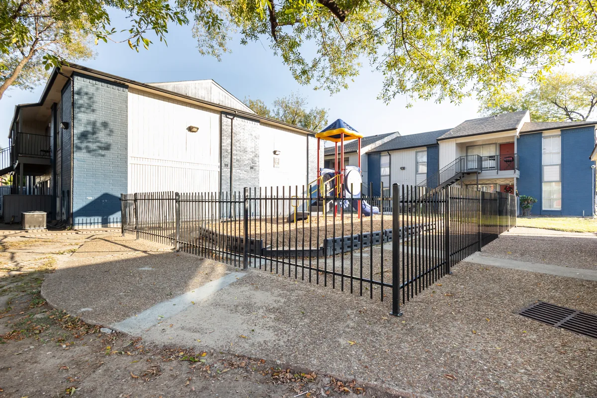 Fenced playground with equipment, surrounded by two residential buildings