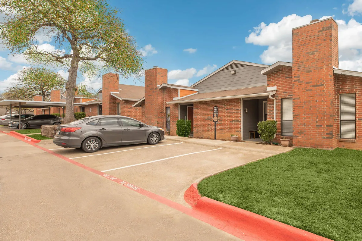 Brick apartment buildings with tall chimneys, mature shade trees, and a landscaped lawn under a blue sky