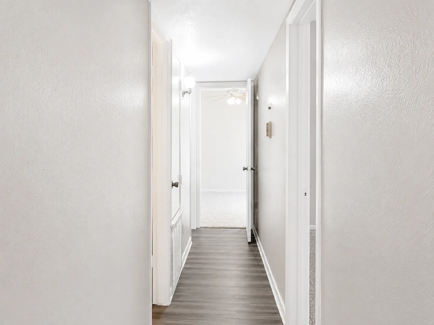 B1 interior hallway with wood-look flooring and panel doors, opening to a carpeted bedroom at the end