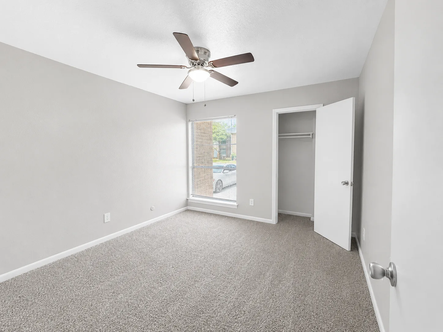 Empty carpeted C1 bedroom with a ceiling fan, a blinded window and an open closet with a hanging rod