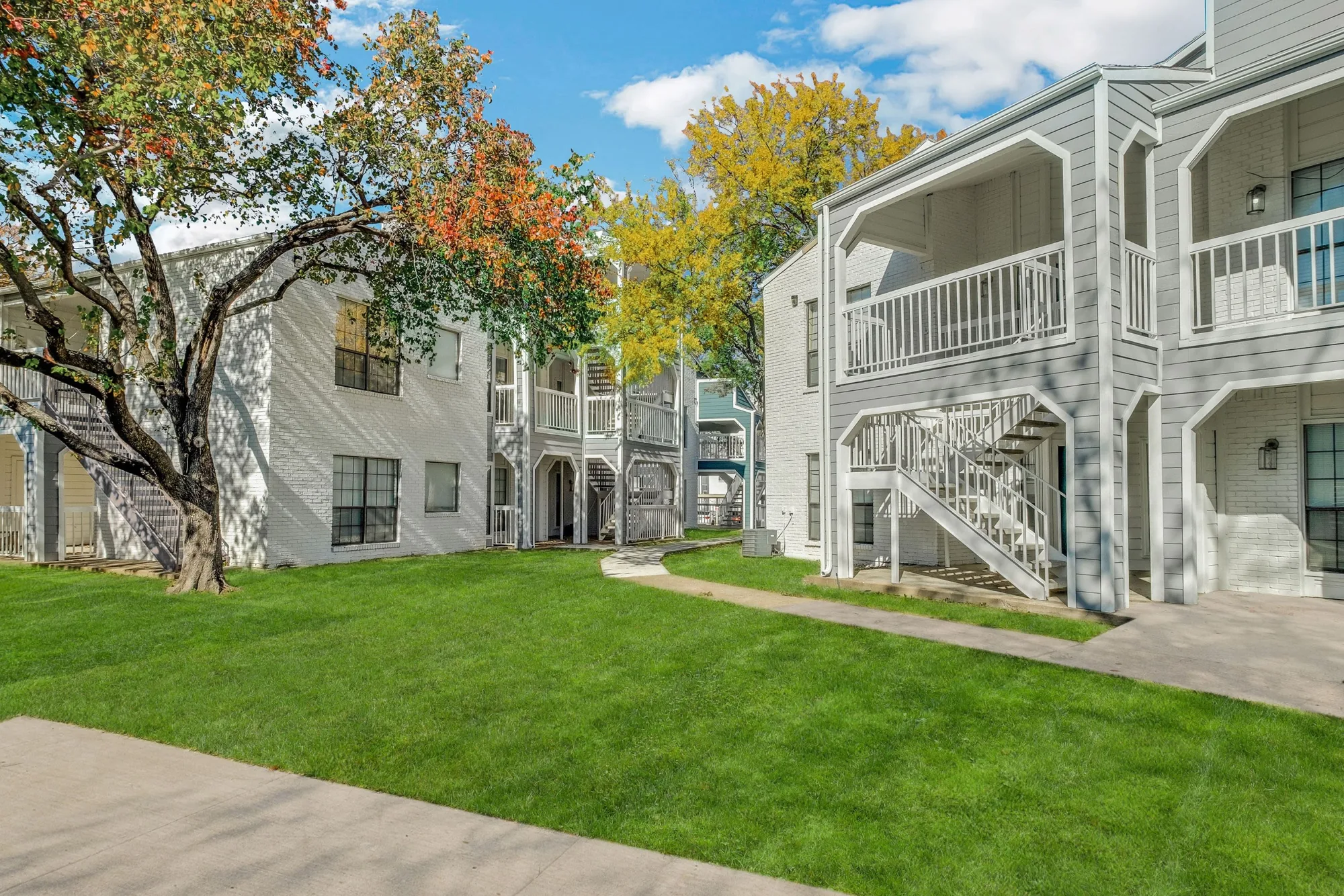 White two-story buildings with balconies and exterior stairs around an open lawn under autumn trees